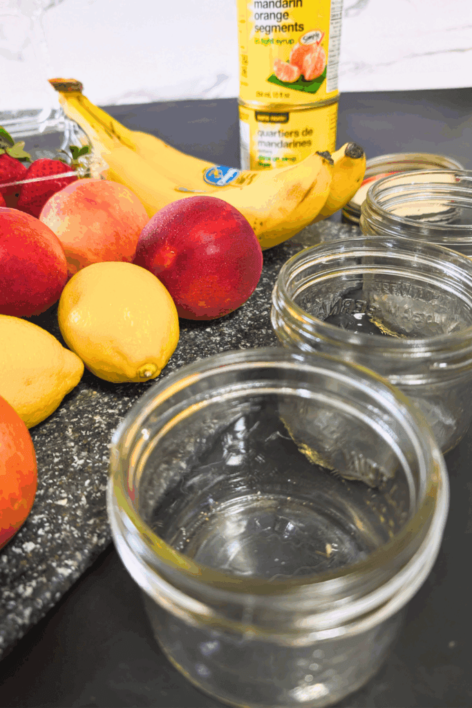 Cut fruit and jar for homemade preserves or jam, fresh strawberries, peaches, lemons, bananas, mandarin orange segments, and glass jars on kitchen counter.