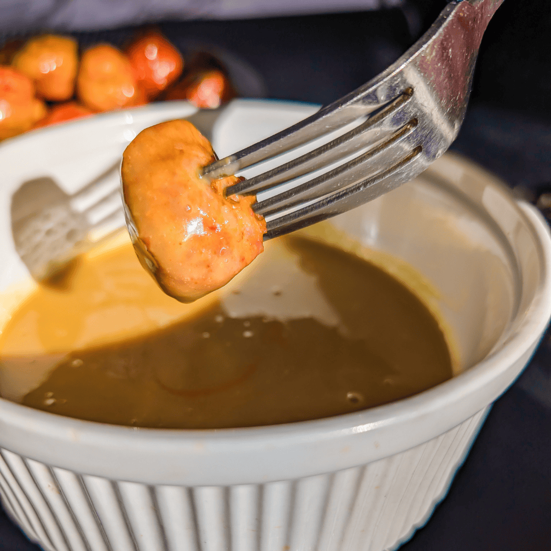Juicy sausage being dipped in rich gravy in a white ramekin.