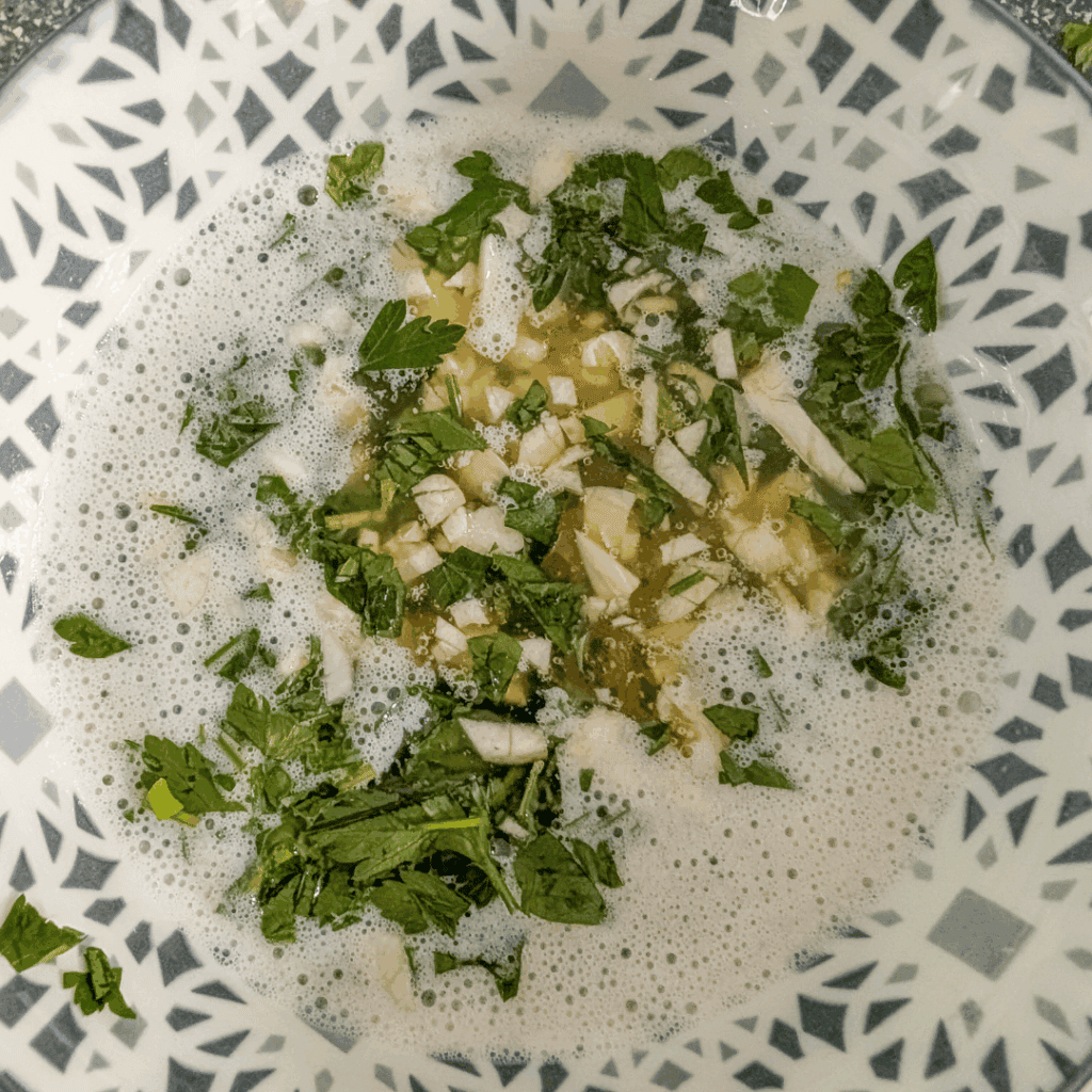 Chopped garlic and fresh herbs in a bowl of bubbling broth for cooking.