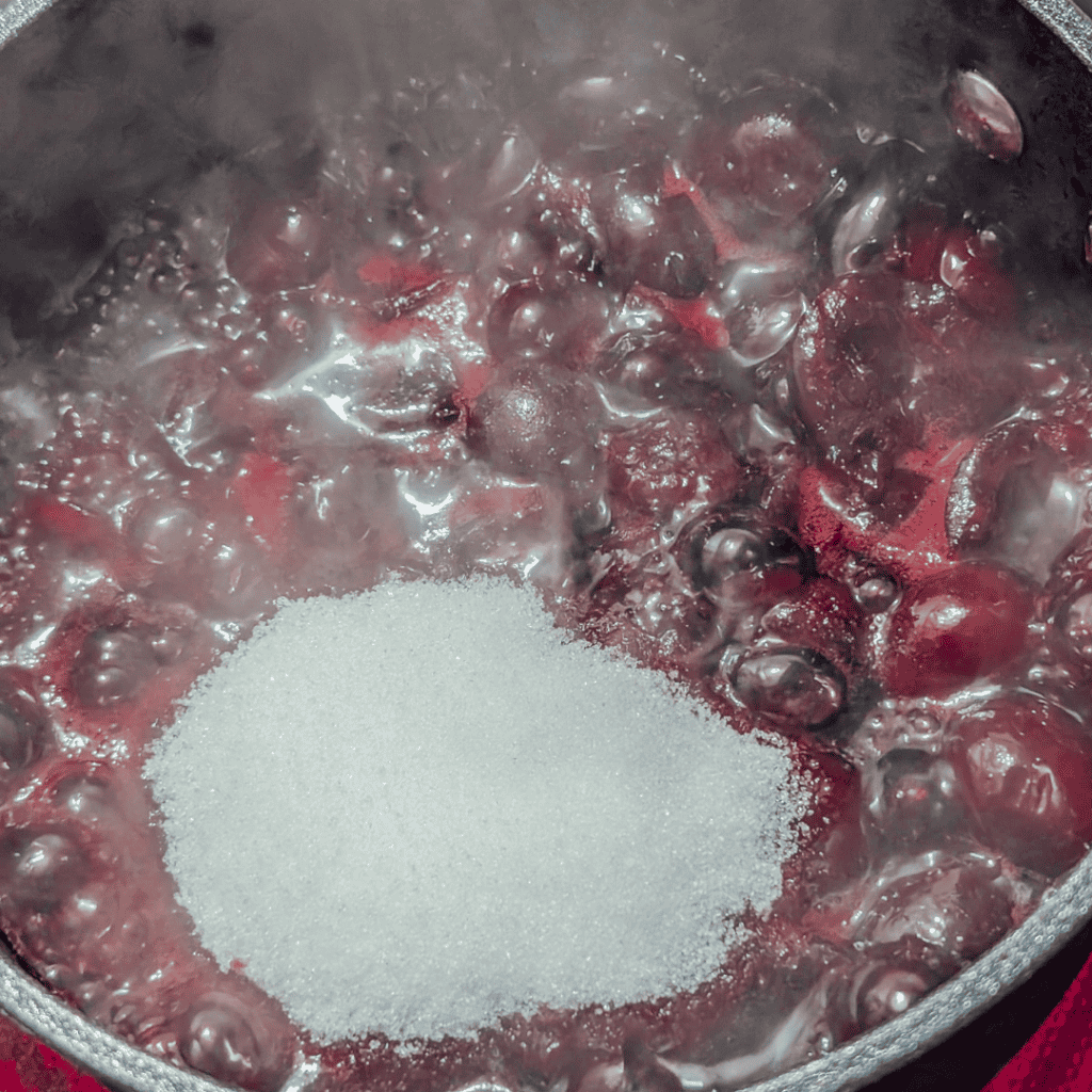 Cherry jam boiling with sugar in a pot, vibrant red cherries and bubbling liquid.