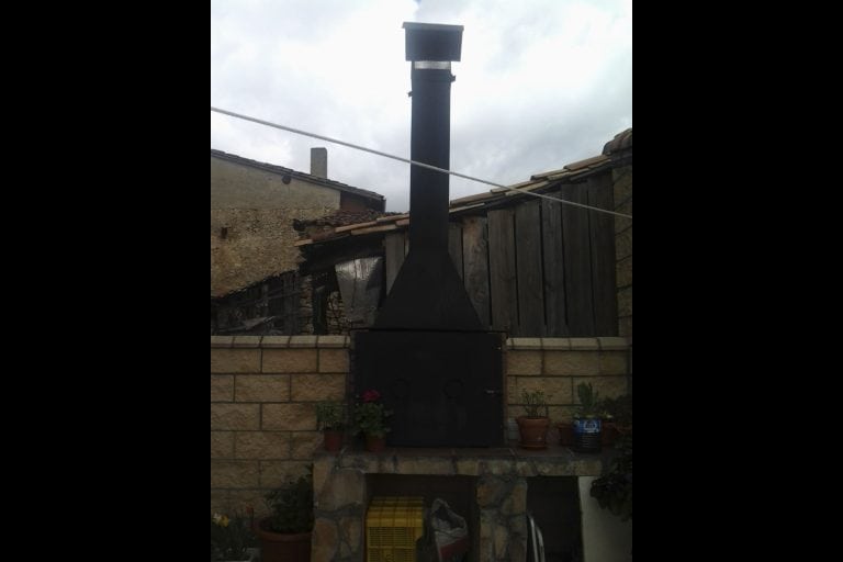 Chimenea exterior de leña negra para calefacción, ideal para jardines, con plantas y estructura de ladrillos y madera.