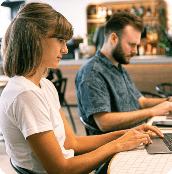 Two people sitting at a table, each working on a laptop in a modern indoor setting.