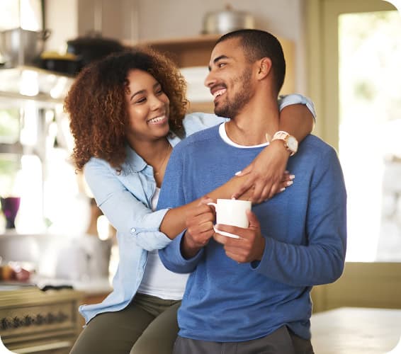 A woman hugs a man from behind in a kitchen while he holds a white mug, both smiling and looking at each other.
