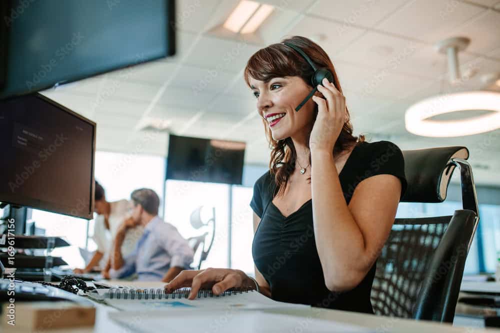 Woman wearing a headset sits at a desk, smiling at her computer monitor, with colleagues in the background of a modern office that manages Dedicated Servers.