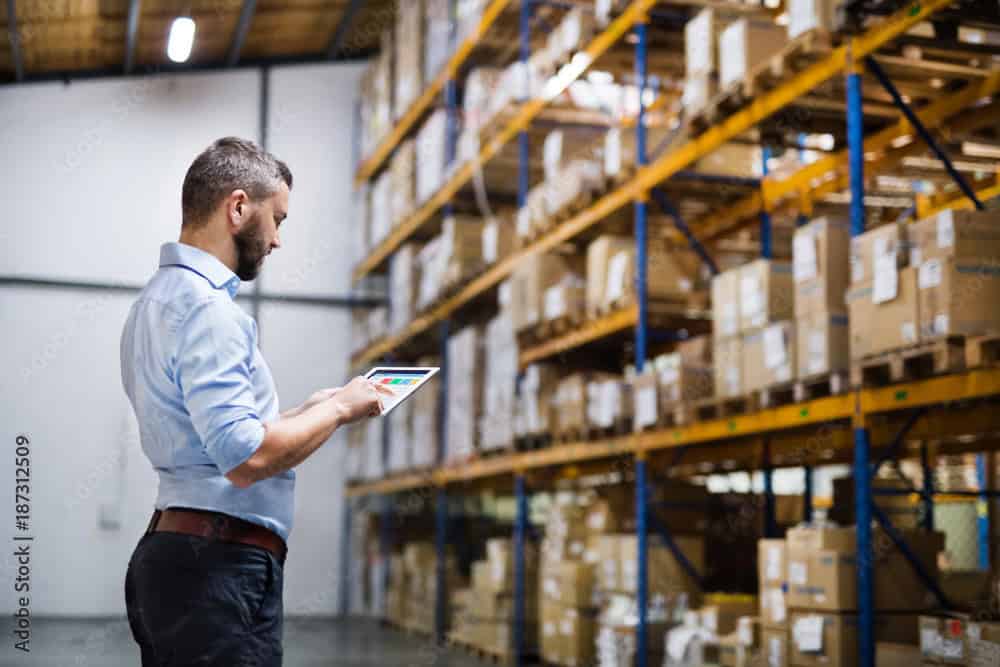 A man standing in a warehouse aisle checks inventory data on a digital tablet, surrounded by shelves filled with stacked cardboard boxes—his workflow powered by efficient dedicated servers for real-time updates.