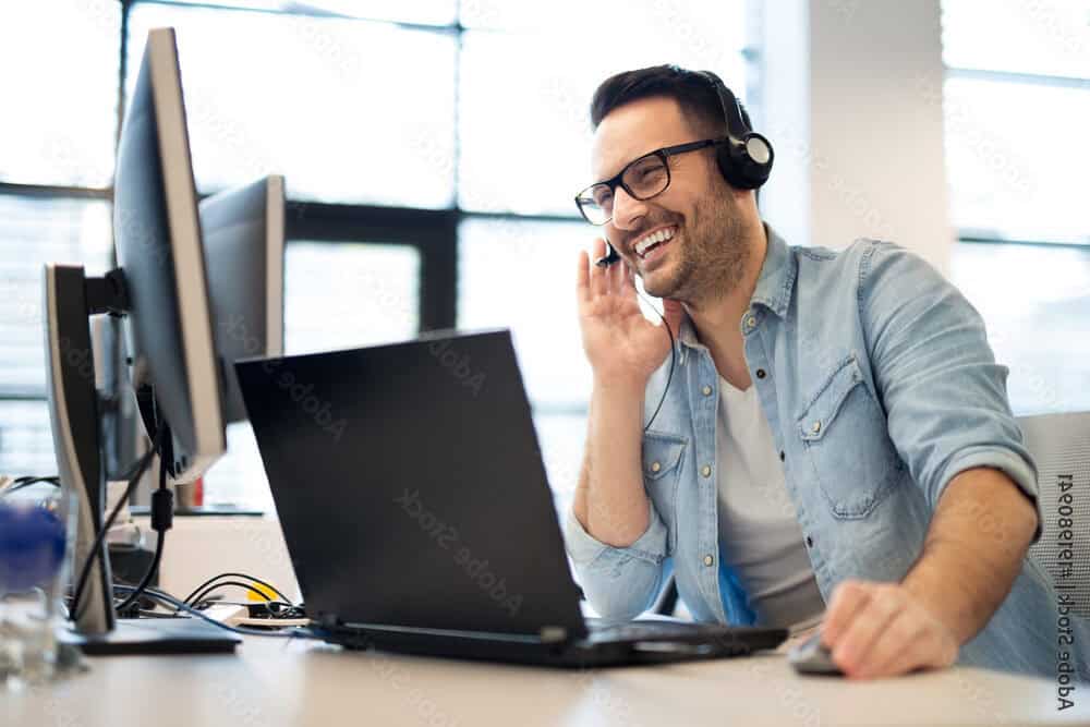 Man wearing glasses and a headset sits at a desk with two monitors and a laptop, smiling while discussing shared hosting solutions during a video or phone call in a bright office setting.