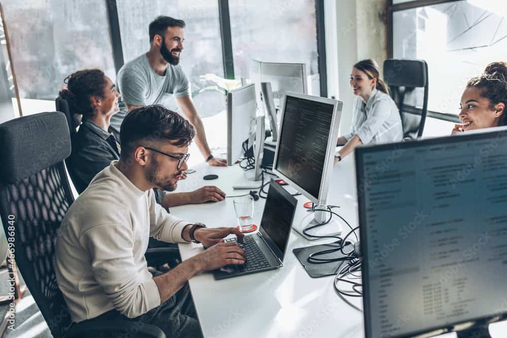 Five people work at desks with computers in a modern office, some collaborating on ASP.NET Hosting solutions while others are focused on their screens.