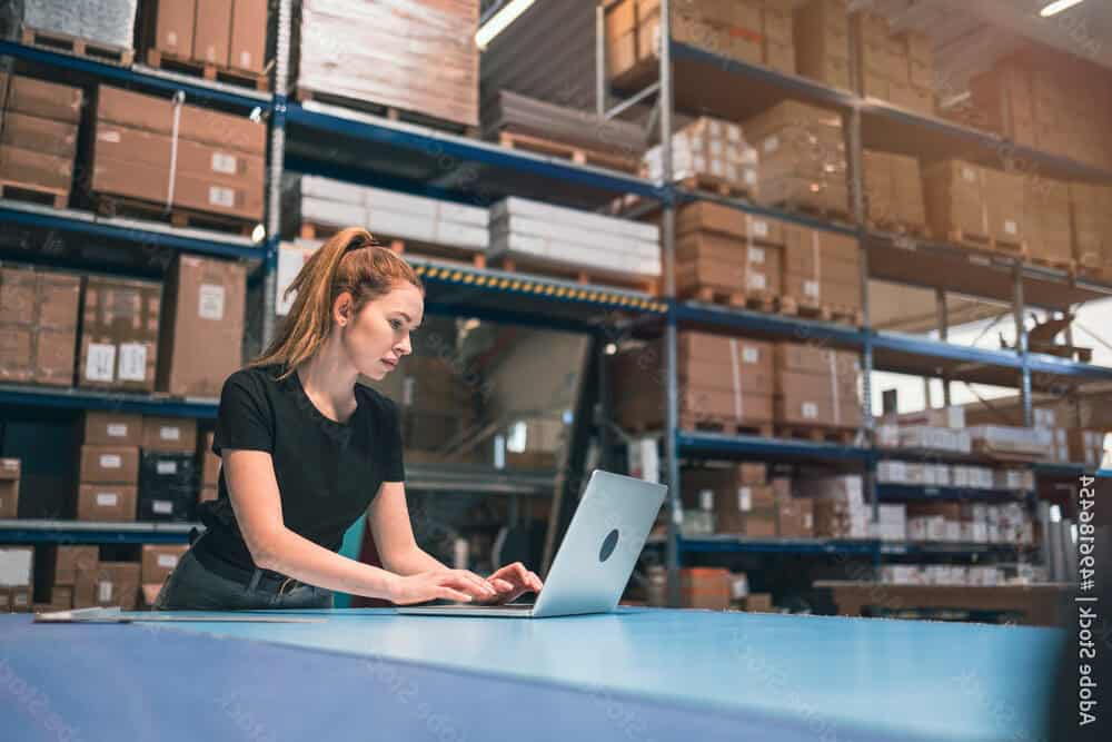 A woman stands at a table in a warehouse, working on a laptop as she manages inventory and coordinates shared hosting logistics with shelves of boxes and packages in the background.