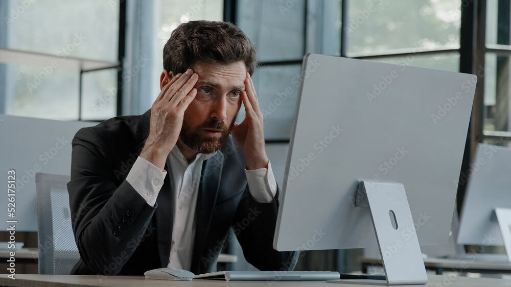 A man in a suit sits at a desk, holding his head in his hands and looking stressed while staring at a computer monitor, possibly facing issues with Dedicated Servers in an office.