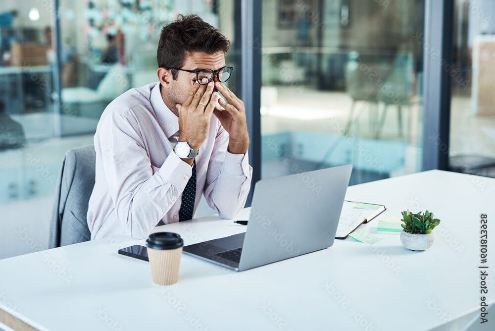A man in business attire sits at a desk with a laptop, coffee cup, and papers, rubbing his eyes while appearing stressed or tired—possibly overwhelmed by shared hosting issues—in an office setting.