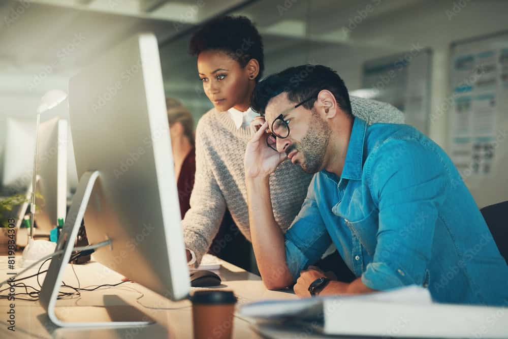 Two colleagues look at a computer screen; one appears stressed with his hand on his forehead, while the other stands beside him observing the monitor, possibly discussing issues related to ColdFusion Hosting.