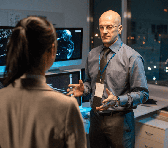 A man in business attire talks to a woman in an office with multiple computer monitors displaying data and maps.
