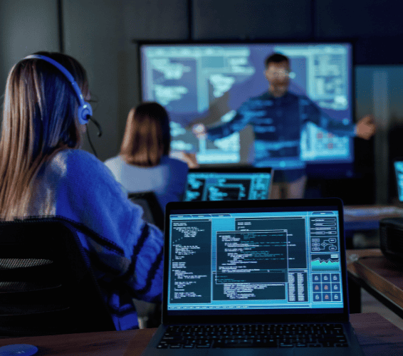 People in a classroom setting attend a lesson on programming; code is displayed on laptops while an instructor presents in front of a projection screen.