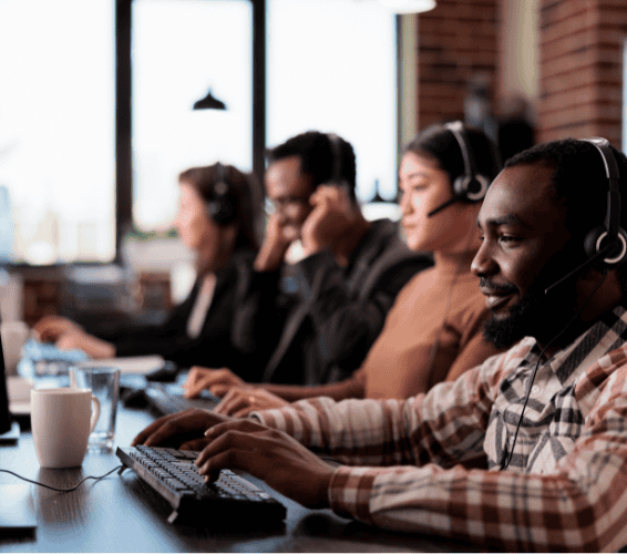 Four people wearing headsets work at computers in a modern office, appearing focused and engaged in customer service or support tasks.