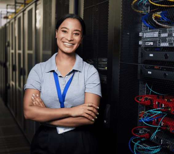 A woman wearing a lanyard stands with arms crossed, smiling, next to server racks in a data center.
