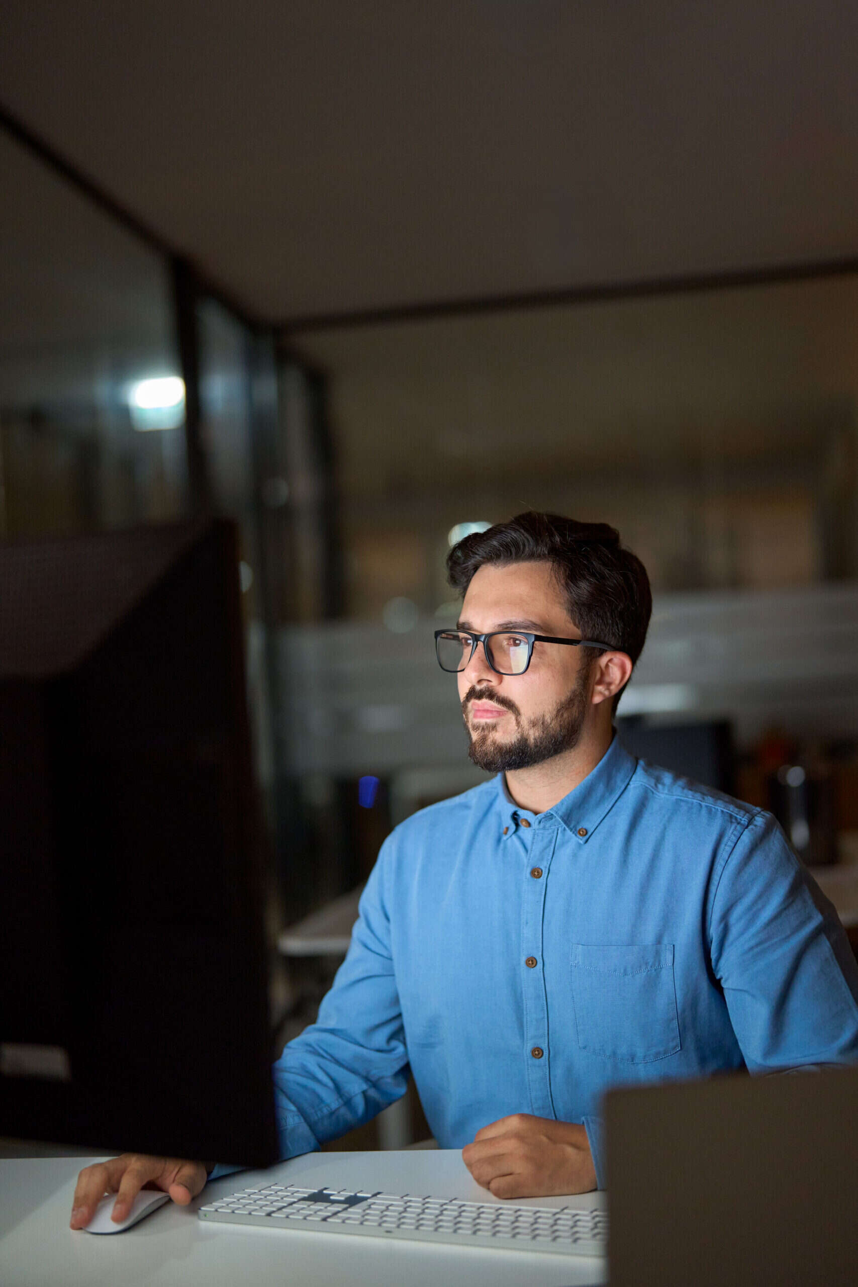 A man wearing glasses and a blue shirt works on a computer at an office desk at night, focusing on configuring WordPress Hosting.