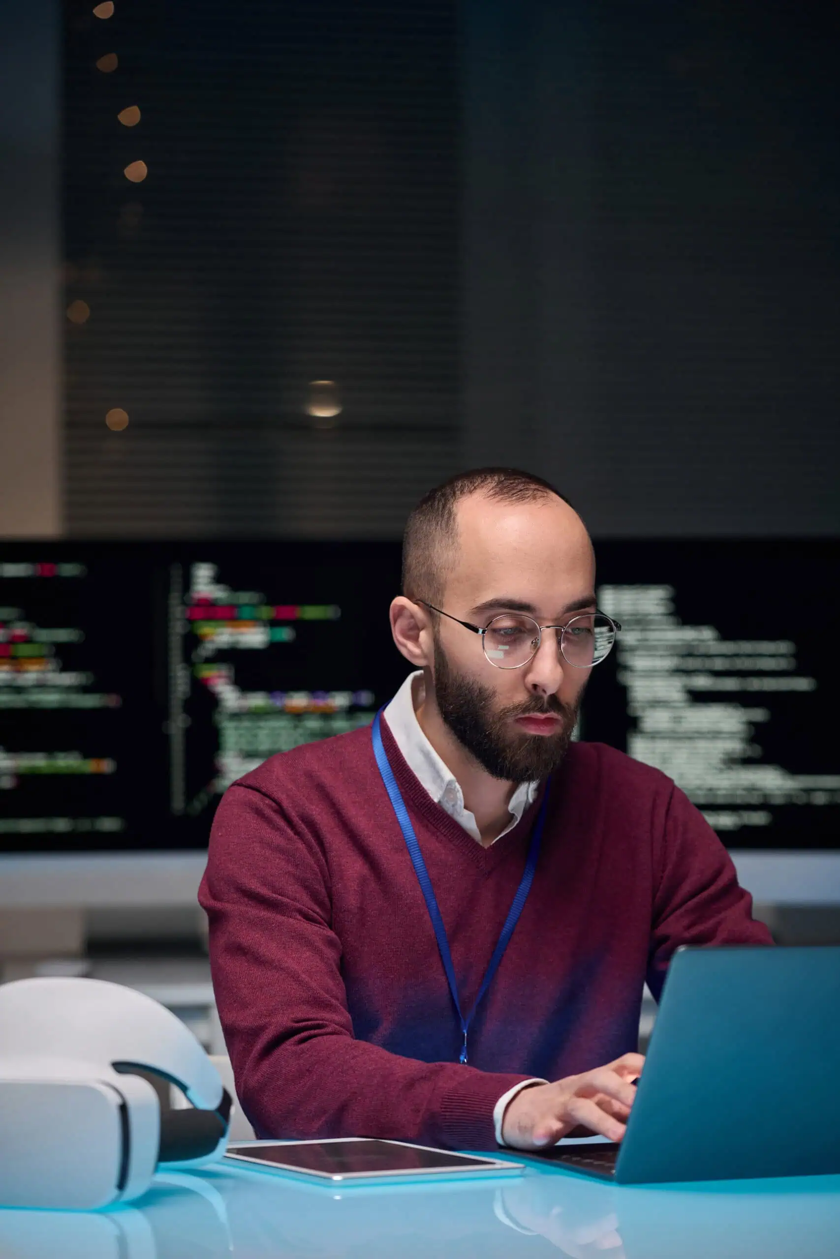 A man with glasses and a beard works on a laptop in an office, with computer monitors displaying code in the background, showcasing the efficiency of Enterprise Hosting Solutions.