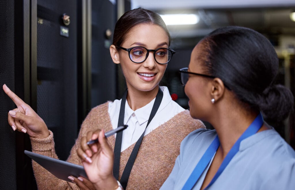 Two women in professional attire discuss MoverEXPRESS solutions while working in a server room; one points at equipment, the other holds a pen and tablet.