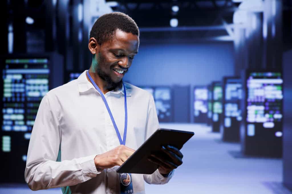 Man in a server room smiling while using a tablet, overseeing maintenance services. He is wearing a white shirt and ID badge, with server racks visible in the background.