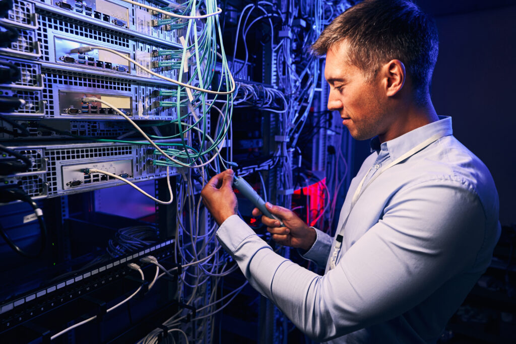 A man in a blue shirt inspects cables and equipment in a server room filled with networking hardware, highlighting the importance of reliable Enterprise Hosting Solutions.