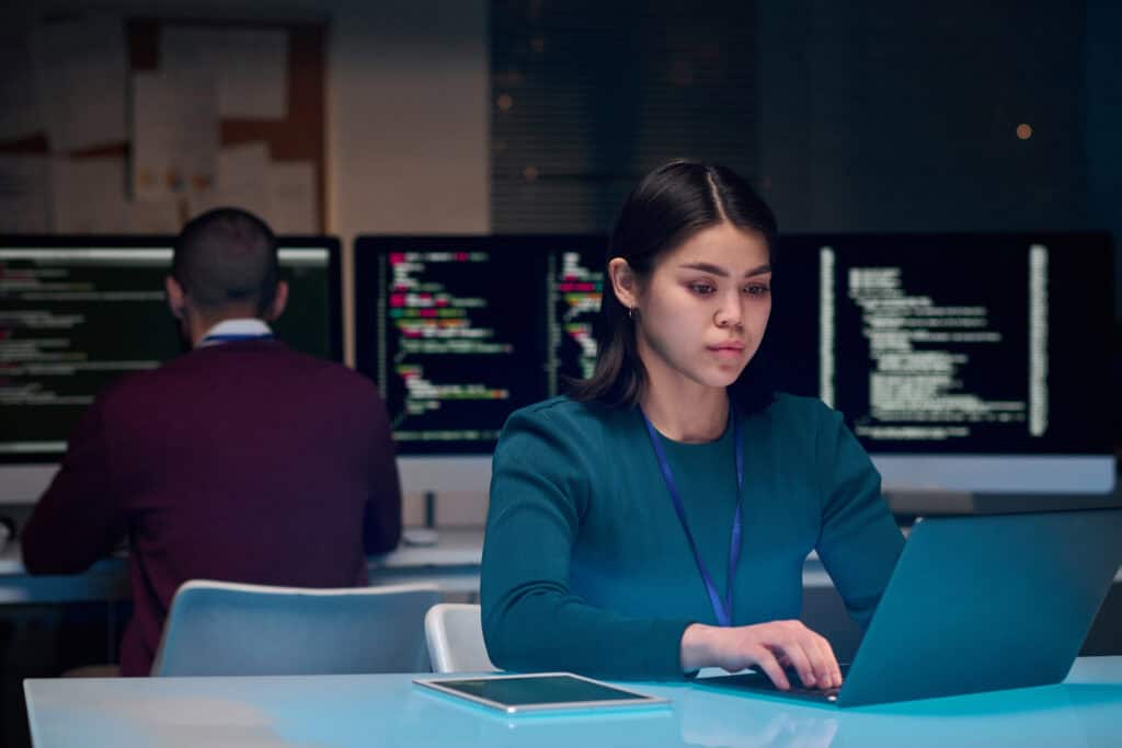 A woman works on a laptop at her desk with a tablet beside her, while a man sits nearby at another desk with computer monitors displaying code—both collaborating on Enterprise Hosting Solutions.