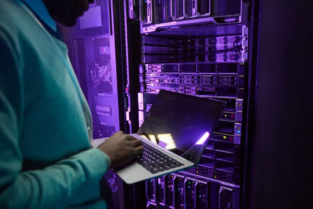 A person uses a laptop while standing in front of server racks in a data center, illuminated by purple lighting.