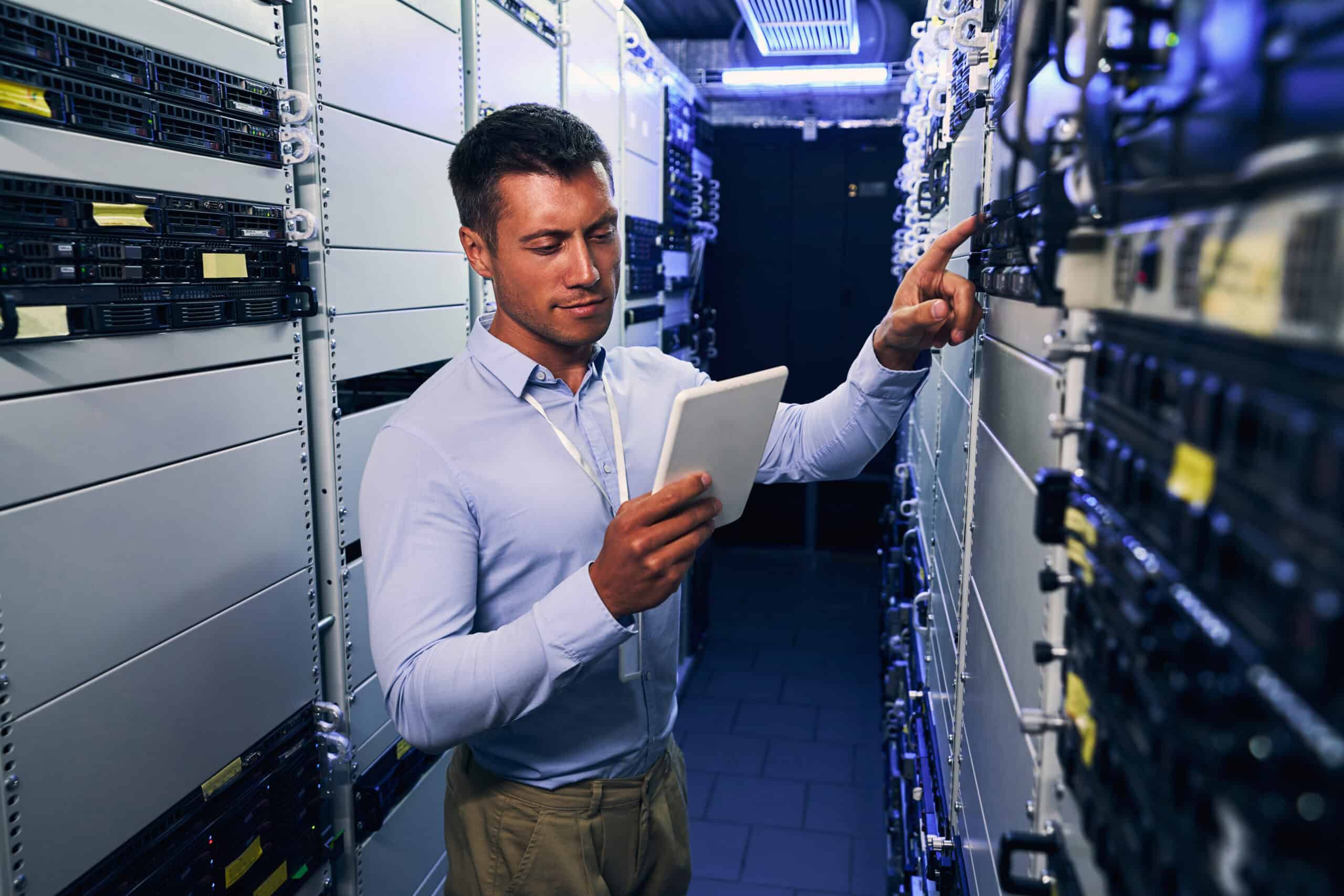 Man in a server room holding a tablet and inspecting server racks, pressing a button on one of the servers—a perfect example of hands-on management for top-tier Enterprise Hosting Solutions.