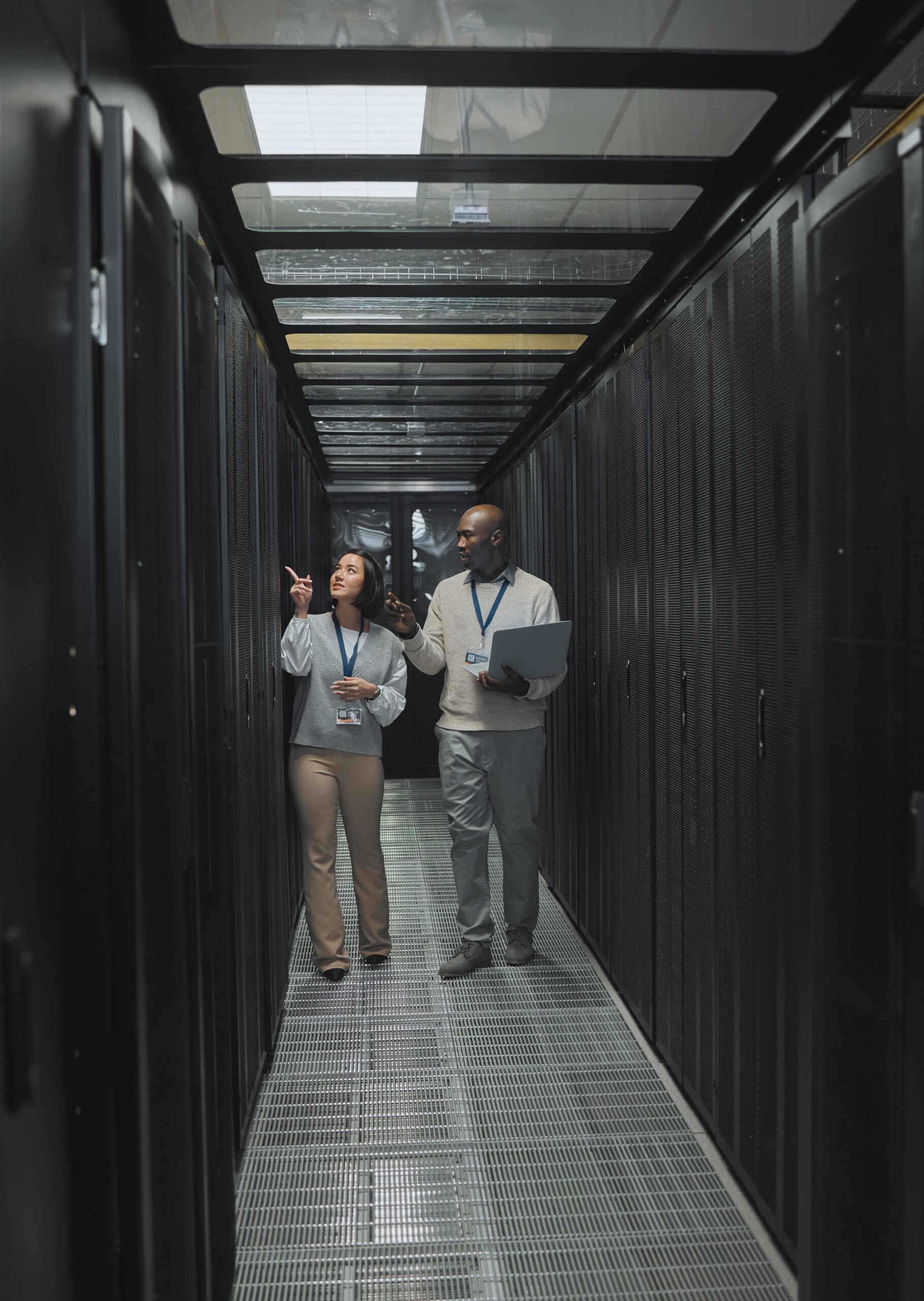 Two people wearing lanyards walk and talk in a server room corridor, surrounded by rows of black server racks hosting Linux VPS solutions.