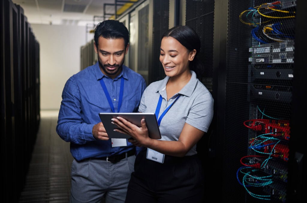 Two IT professionals stand in a server room, looking at a tablet together, with server racks and network cables visible in the background.