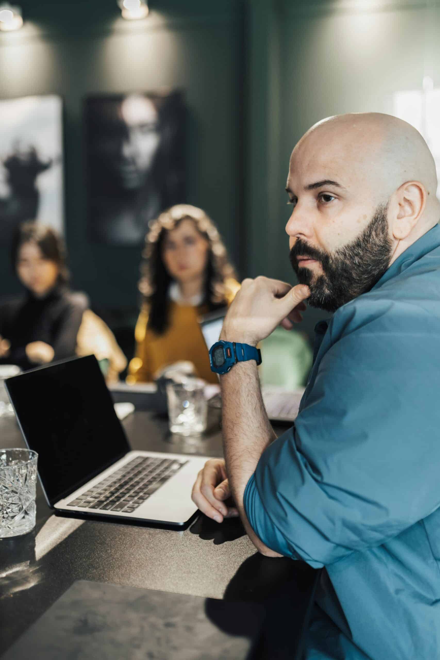 A man with a beard sits at a table with a laptop and glass of water, looking to the side, perhaps discussing ASP.NET hosting. Two women are seated in the blurred background.