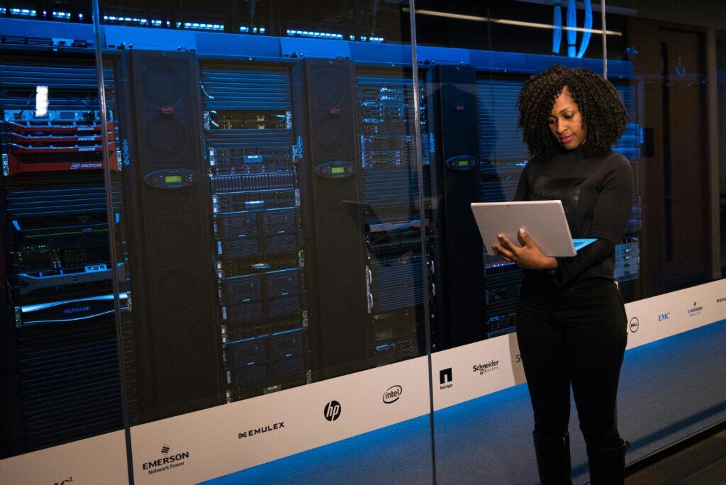 A woman stands in front of server racks, working on a laptop in a data center environment, ensuring reliable ASP.NET Hosting for seamless web application performance.