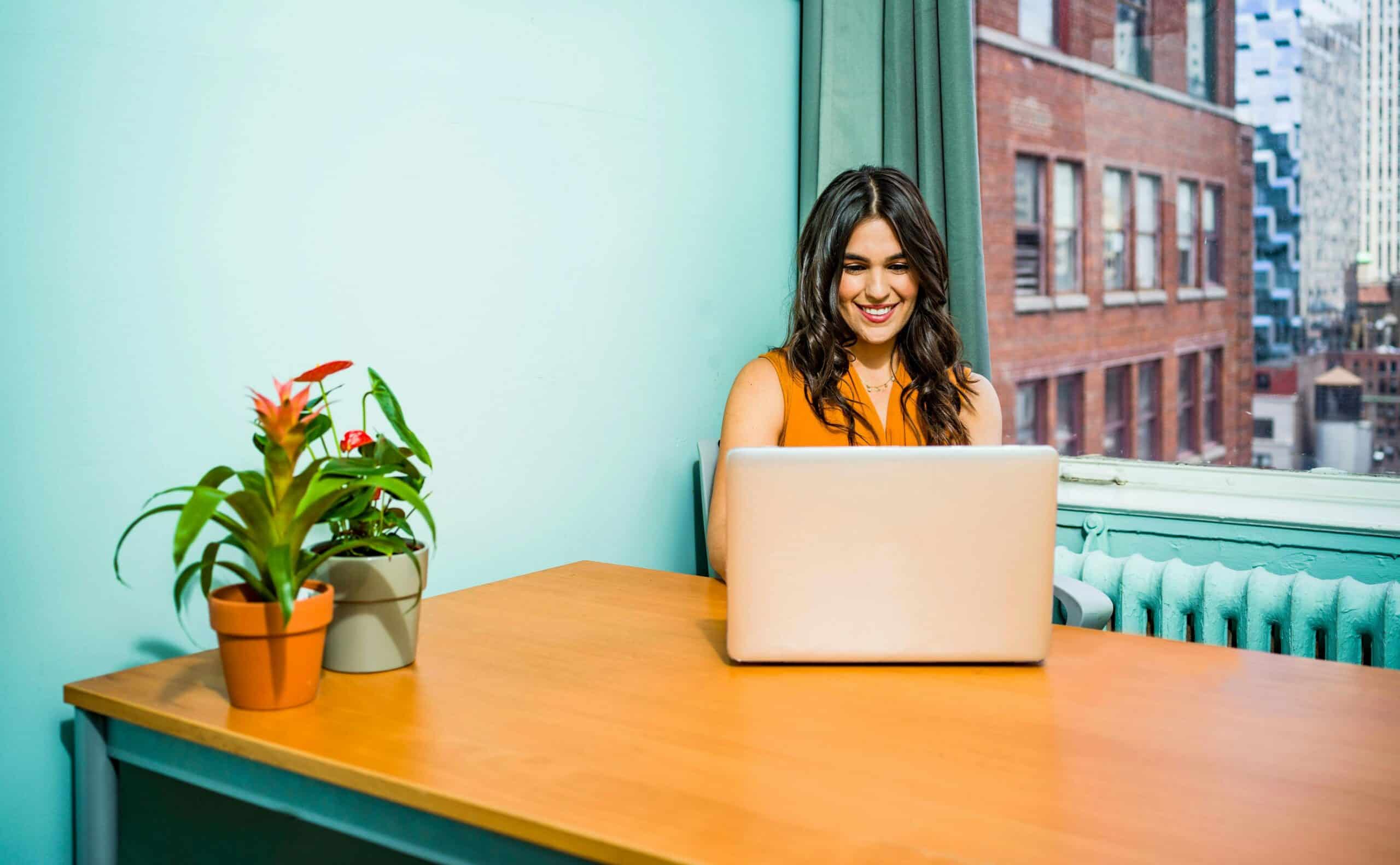 A woman sits at a wooden desk with a laptop and two potted plants, smiling, in a brightly lit office with large windows overlooking city buildings—her shared hosting dashboard open on the screen.