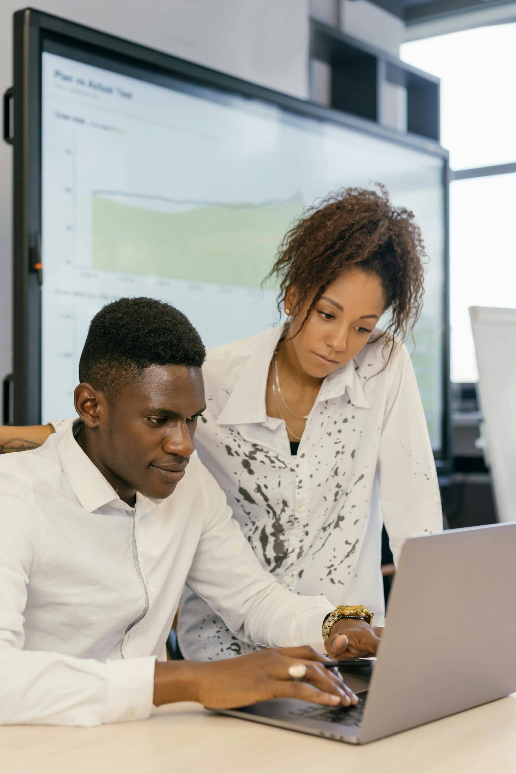 Two people in white shirts are looking at a laptop screen together in an office, discussing ColdFusion Hosting, with a large monitor displaying a graph in the background.