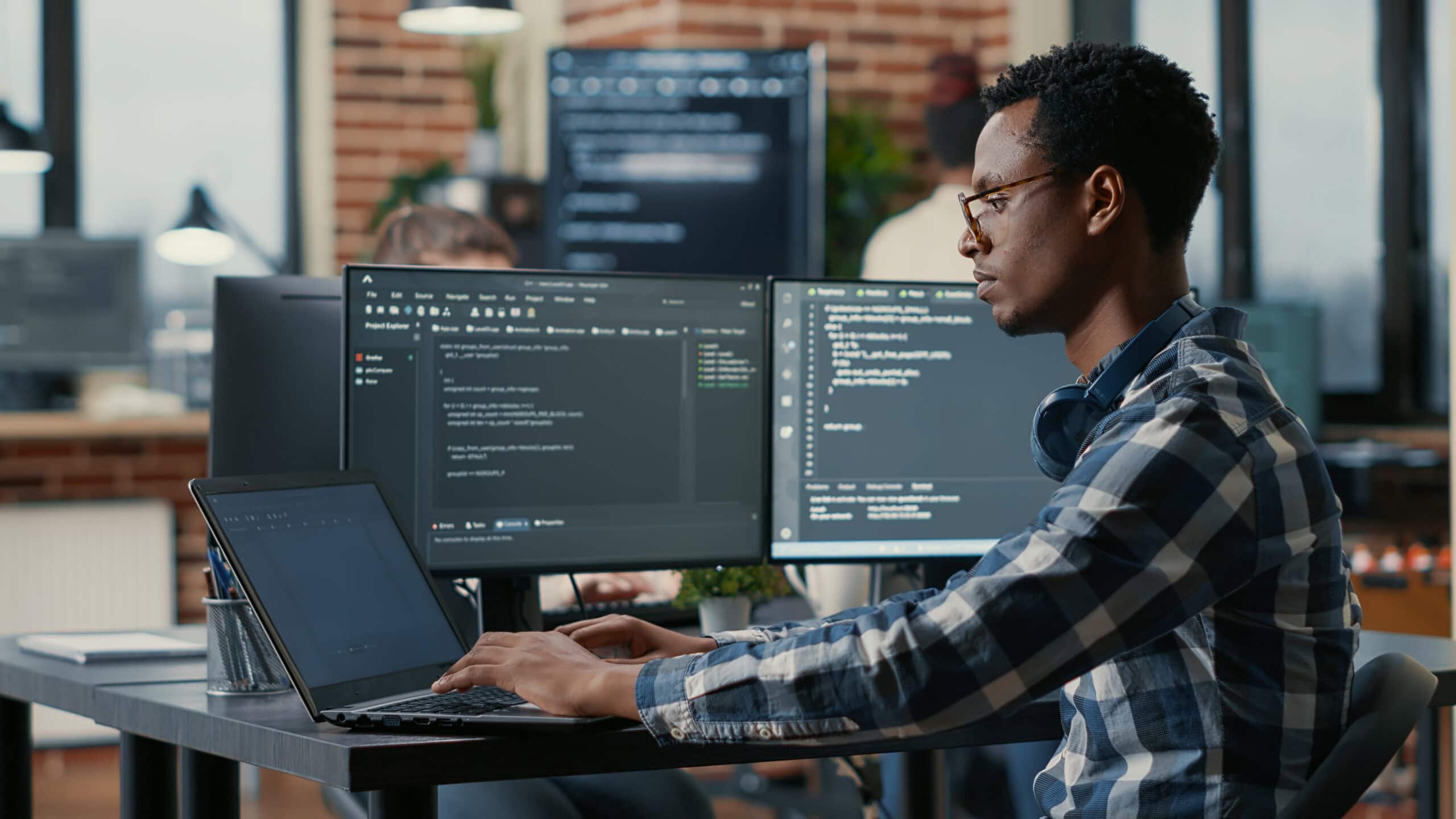 A person in glasses works on a laptop and two external monitors displaying code, focused on WordPress Hosting tasks, seated at a desk in a modern office environment.