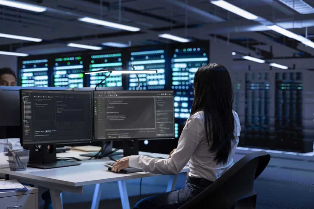 A person sits at a desk with two monitors displaying code in a modern office, with server racks and data center equipment visible in the background.