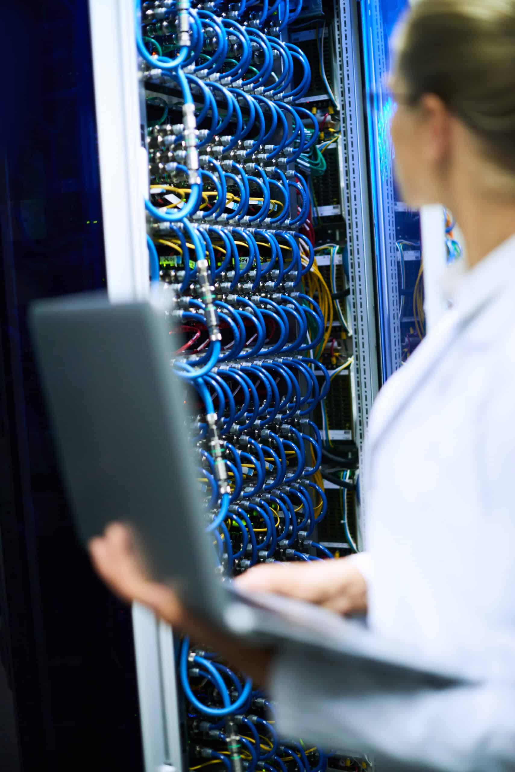 A person holding a laptop stands in front of a server rack filled with network cables and hardware, illustrating the backbone of Enterprise Hosting Solutions in a modern data center.