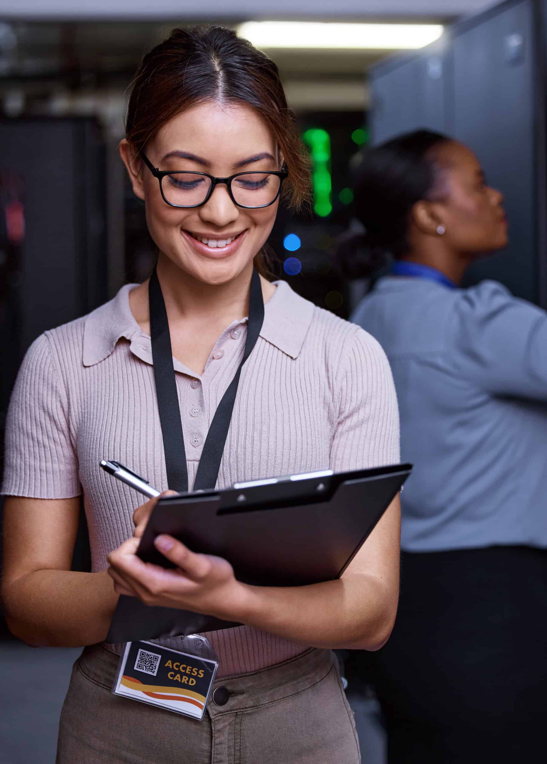 Woman wearing glasses and an access card writes on a clipboard in a server room with dedicated servers, while another woman works in the background.