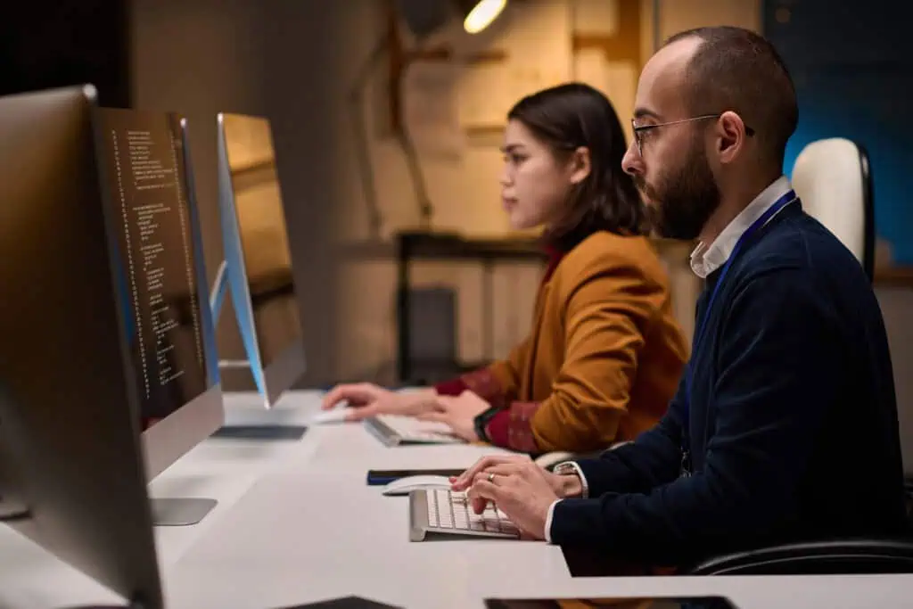 Two people sit at a desk working on desktop computers in an office, typing and looking at their monitors displaying code as they collaborate on Enterprise Hosting Solutions.