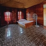Cozy wooden bedroom with natural decor, featuring a comfortable bed, windows with curtains, and a standing fan.