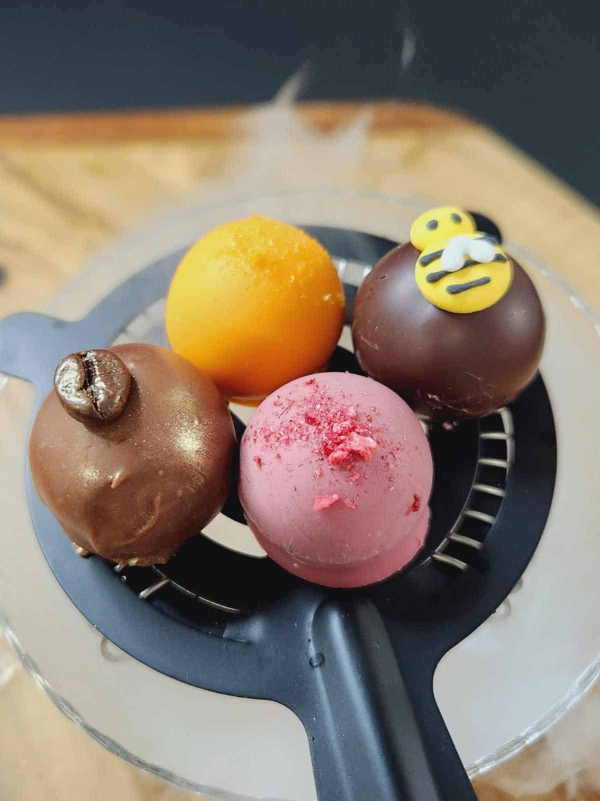 Four colorful chocolate truffles sit on a circular black tray, including one yellow, one pink, and two brown truffles, with unique decorative toppings on each.