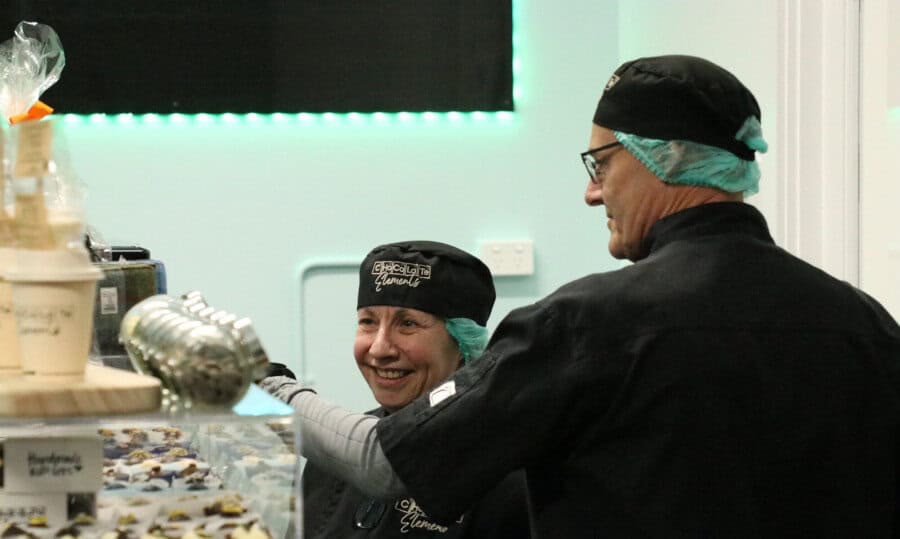 Two people wearing black uniforms, hats, and hairnets work together behind a counter with various treats and jars in a brightly lit, modern shop. One person is smiling, and both appear focused on their tasks.