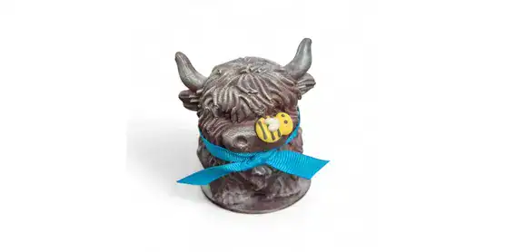 A small figurine of a shaggy cow with horns, wearing a blue ribbon and a tiny bee charm around its nose, on a white background.