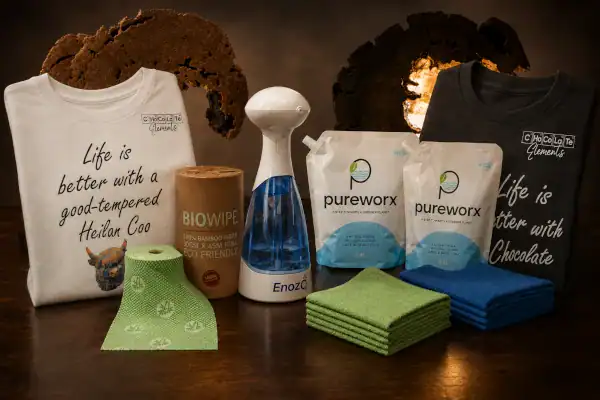 T-shirts, reusable cloths, paper towels, cleaning spray, and cleaning wipes sit on a table with a brown, textured backdrop and chocolate-themed decor in the background.