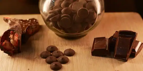 A glass jar of chocolate chips, a chocolate bar broken into pieces, and a chocolate-covered treat sit on a wooden surface.