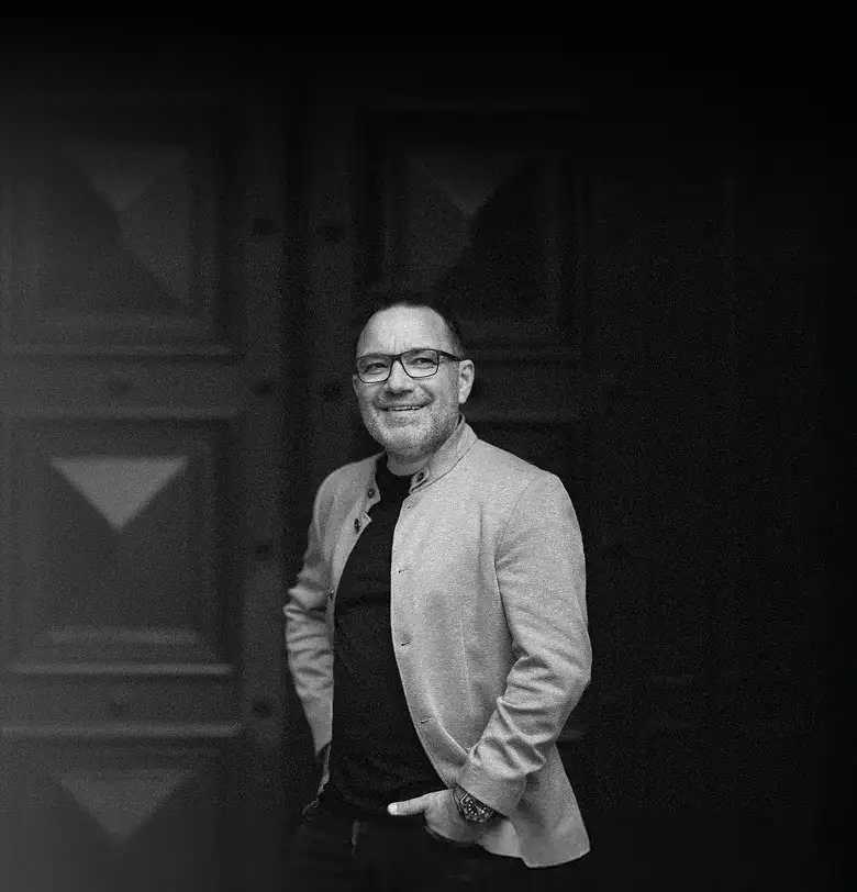 Christian Gröger steht lächelnd vor einer Tür, trägt Brille und helles Sakko in schwarz weiß