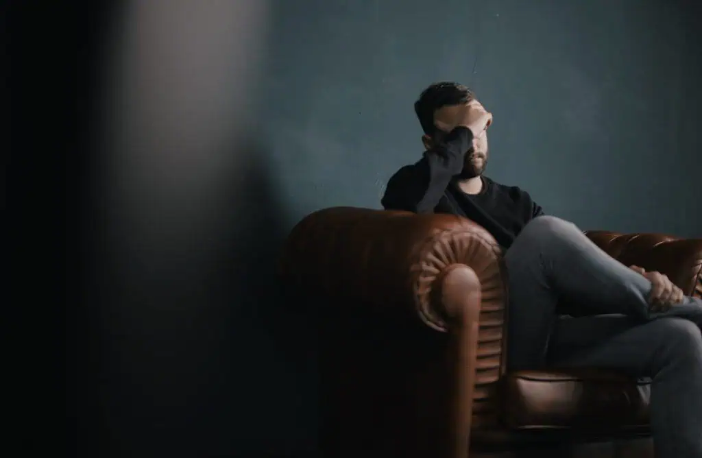 Frustrated man sitting on a brown leather sofa, holding his forehead with a dark background, representing stress or financial concerns related to investing in Bitcoin and cryptocurrency.