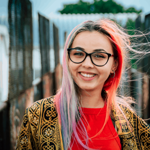 Bright young woman with colorful hair and glasses smiling outdoors, showcasing modern fashion and vibrant personality for Chrysalis BTC SEO optimized image.