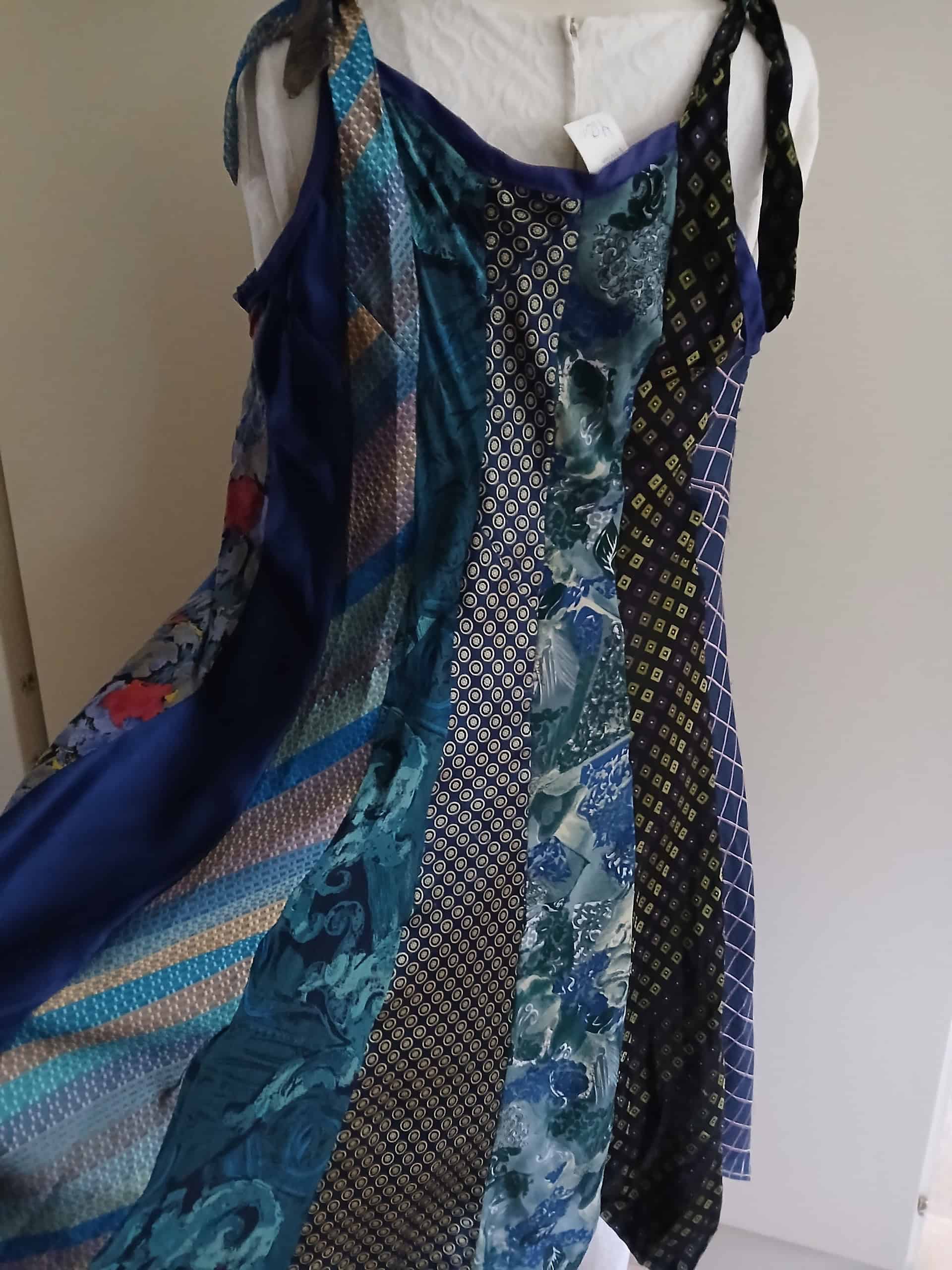 Ljusblå och mörkblå patchworkklänning med olika mönster och texturer, hängande på en person mot en neutral vägg.