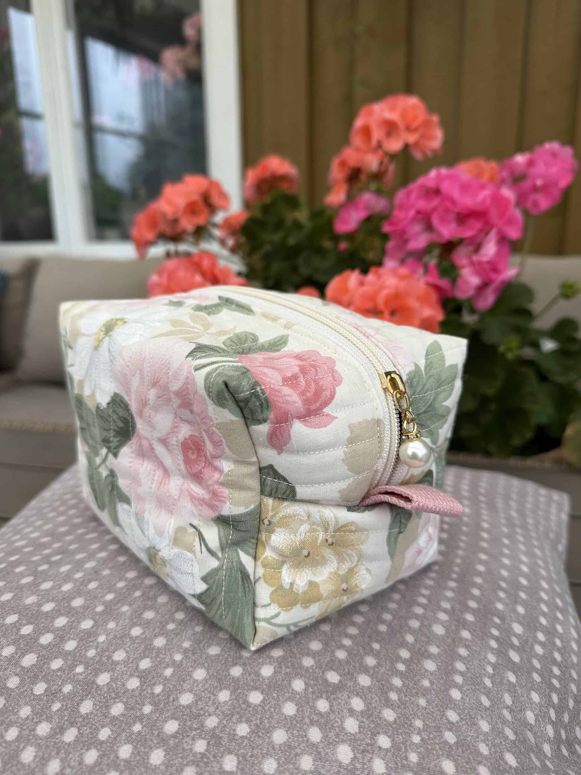 Elegant blommig necessär i tyg med rosa och gröna nyanser, perfekt för förvaring av småsaker, placerad på ett polkadot-tygbord mot en bakgrund av fuchsia- och aprikosfärgade pelargoner.