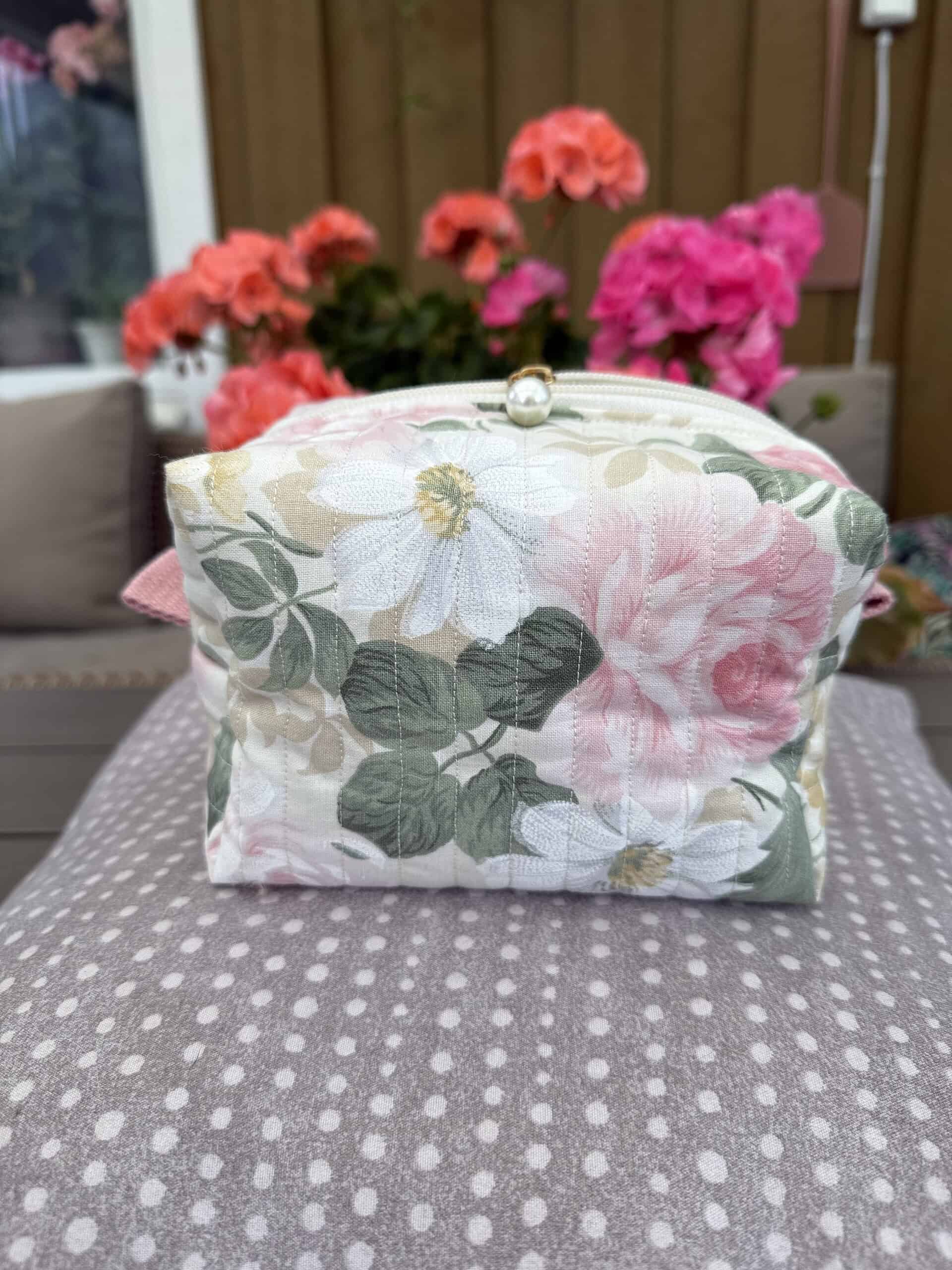 Högupplöst tygväska med blommönster framför en bakgrund av rosa geraniums i trädgård. Perfekt som handväska eller smyckesförvaring, designad av CircleSign.